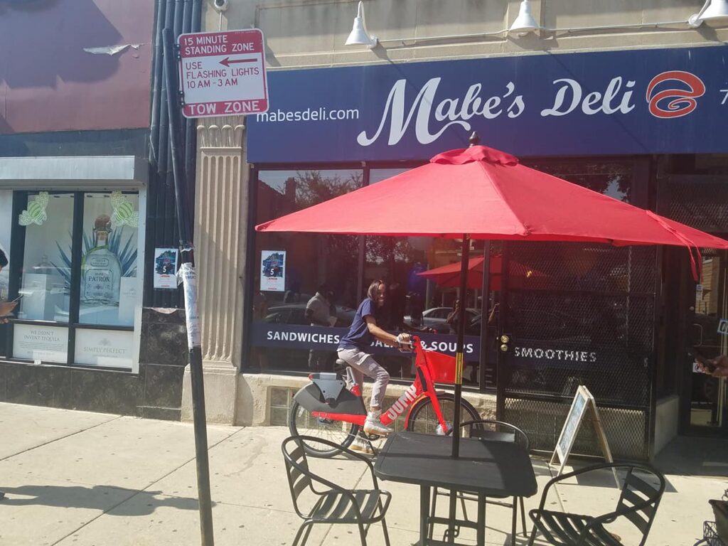 Photo of Mabe's Deli with a red umbrella by outdoor seating on a city sidewalk